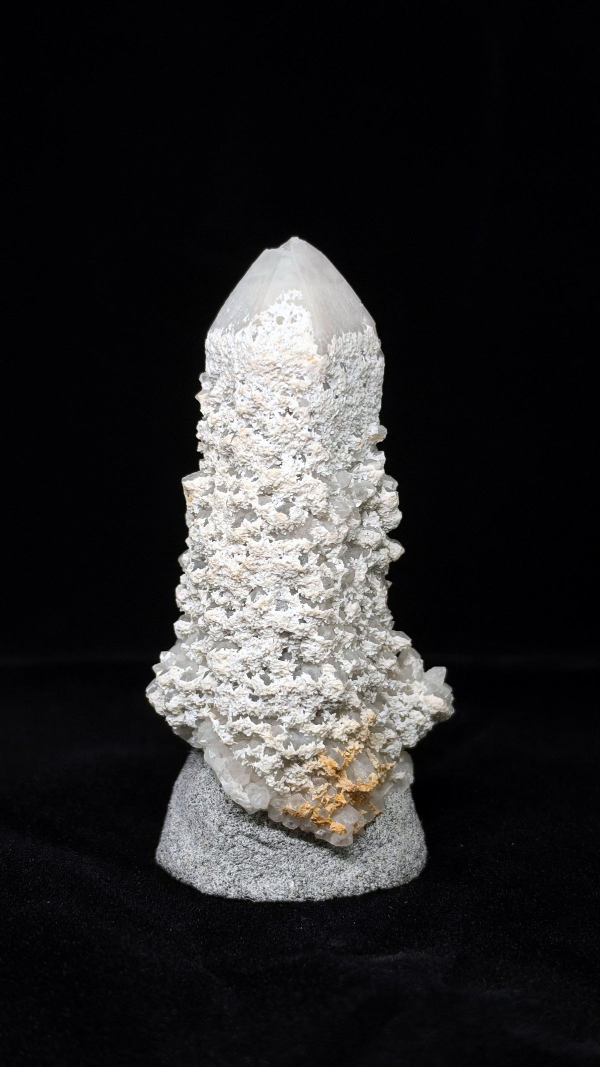 乳白色尖頂標本立於灰色展示座,表面密布白色細碎紋理,右下方有黃褐色斑塊。
