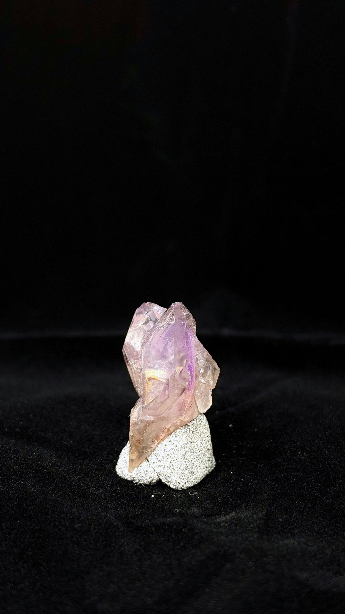晶體的窄側視角,前後尖端交錯,紫色帶穿過透光面,下緣斜向延伸至灰白底座前方。