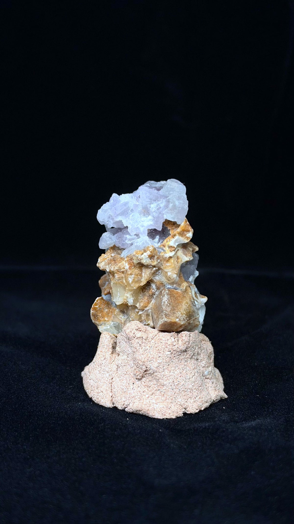 較窄視角下的直立標本，頂部淺紫晶體向左伸展，下方褐色塊面突出，置於粉米色底座上。